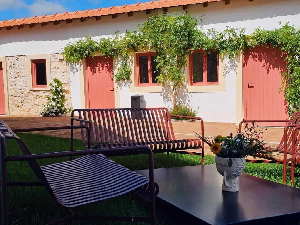 En terrasse med metalstole og et bord, en hvid bygning med røde døre og vinduer, samt grønt frodigt vegetation.