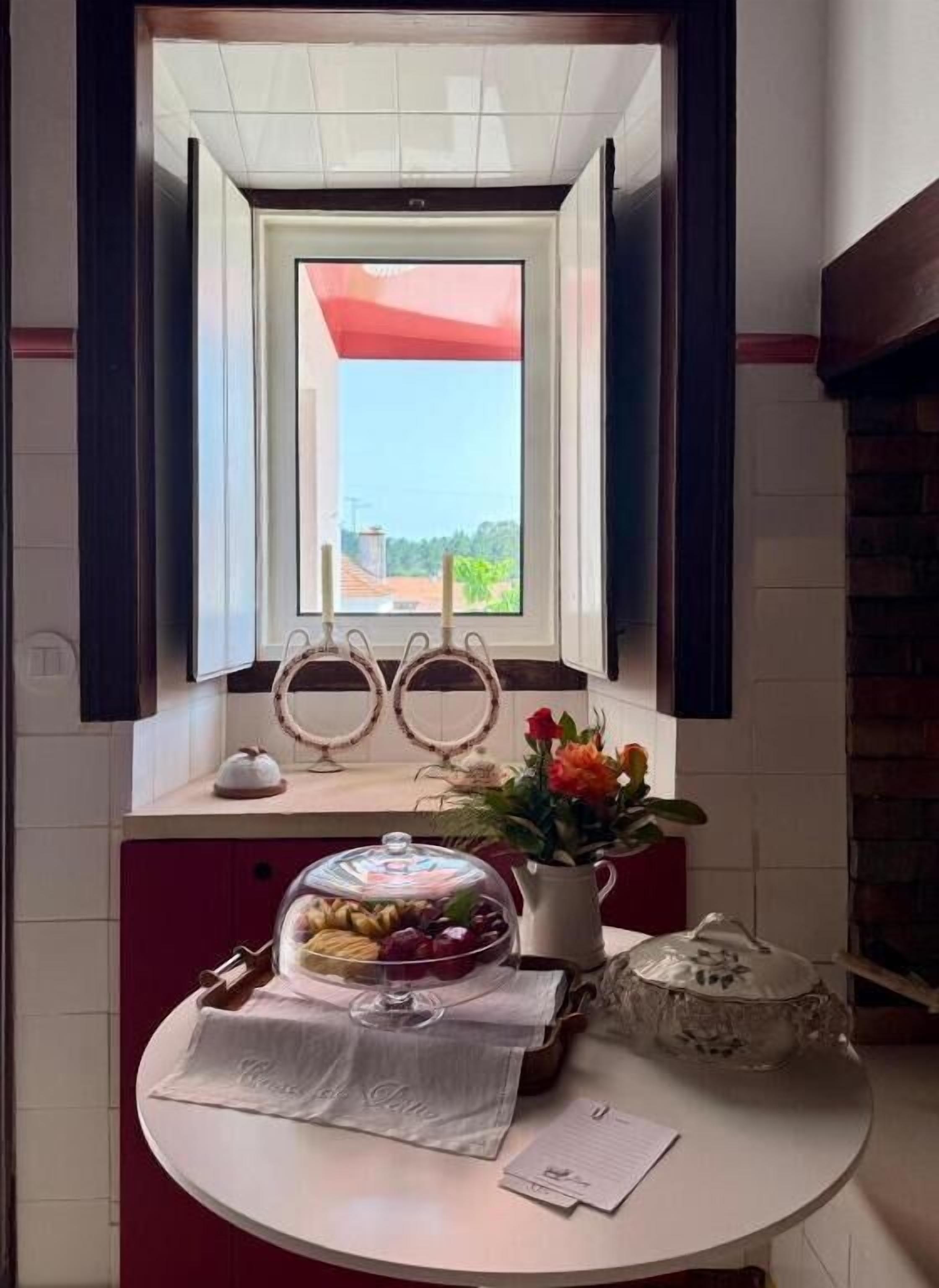 A room with a window, a table with a glass dome, a vase with flowers, and a decorative item on the windowsill.