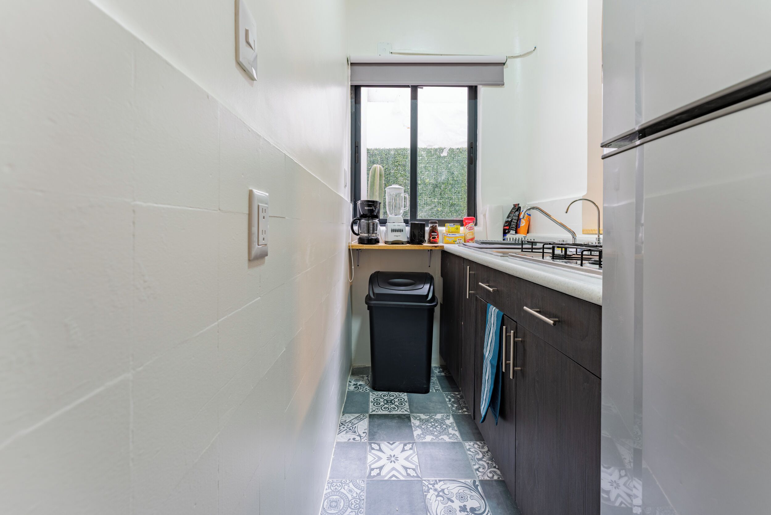 A narrow kitchen with a window, a trash bin, a coffee maker, and a blender on the counter.