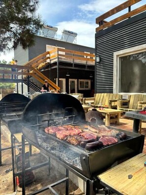 A barbecue grill with multiple meats cooking, a wooden deck, and a building with a window and a staircase. - Diez Pinos (Punta del Este)