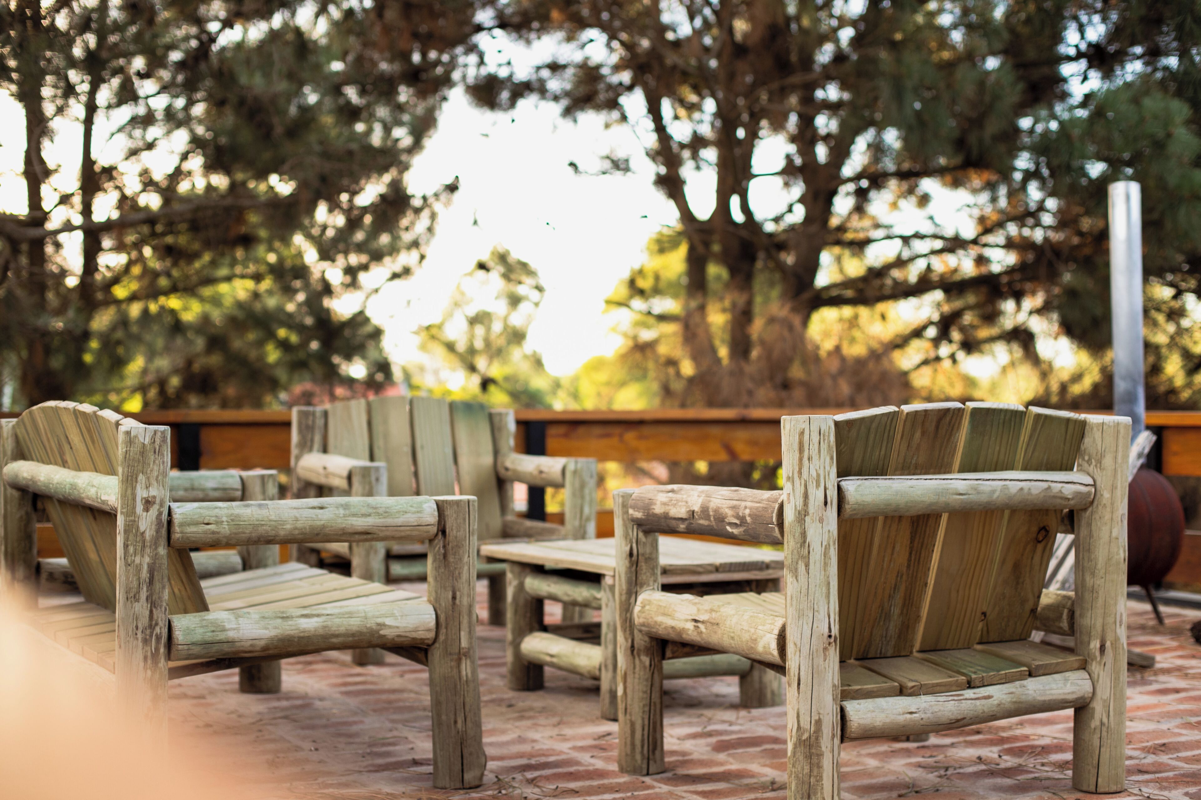 Outdoor wooden furniture arranged on a brick patio with trees in the background.