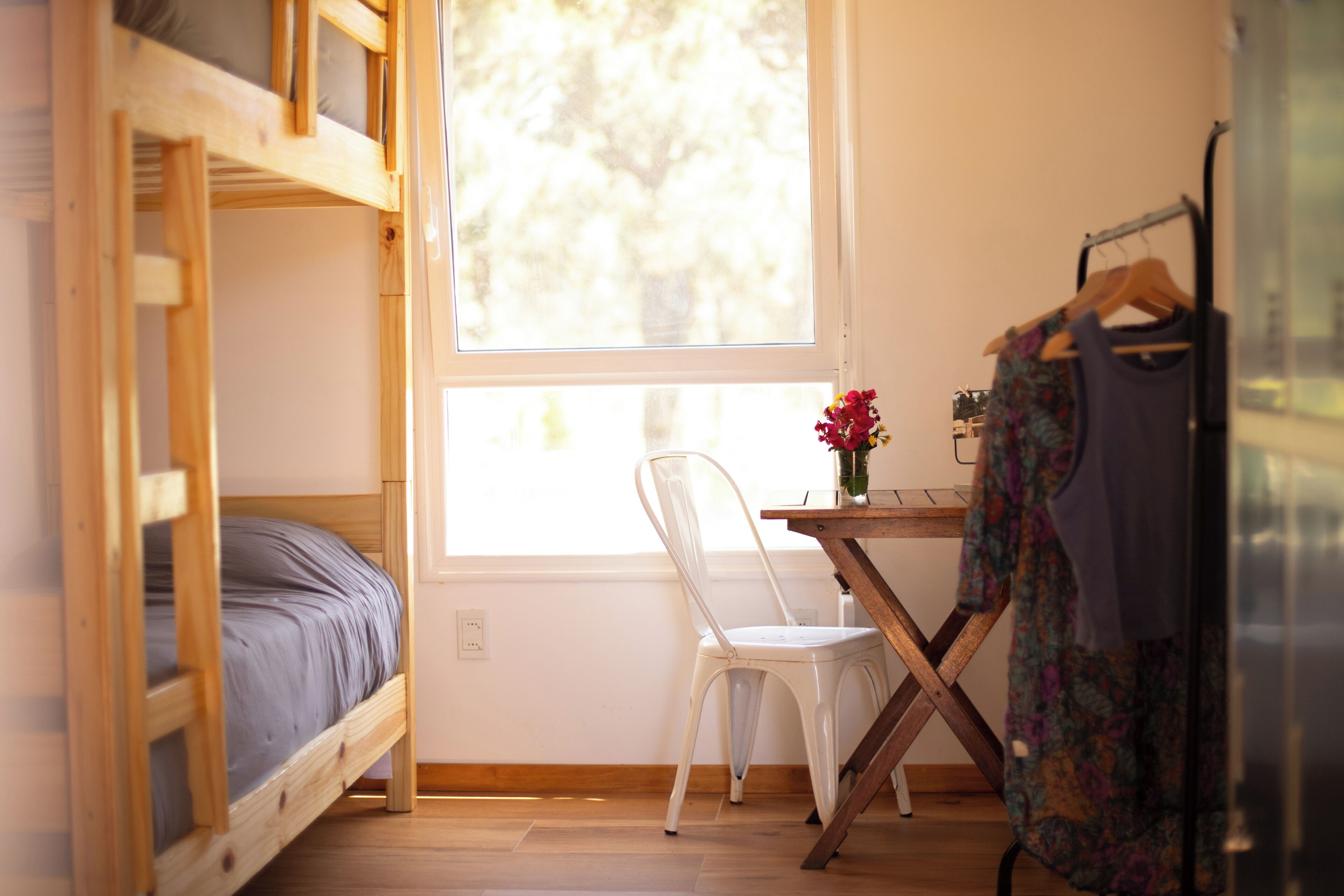 A room with a bunk bed, a white chair, a table with a vase, and a hanging garment.