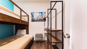Une petite pièce comprenant un lit superposé, un petit bureau, une chaise, une table de chevet, une étagère fixée au mur et une porte.
