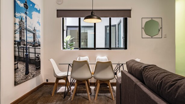 Un espace repas comprenant une table, quatre chaises, un luminaire suspendu, une grande fenêtre et une œuvre d’art accrochée au mur.