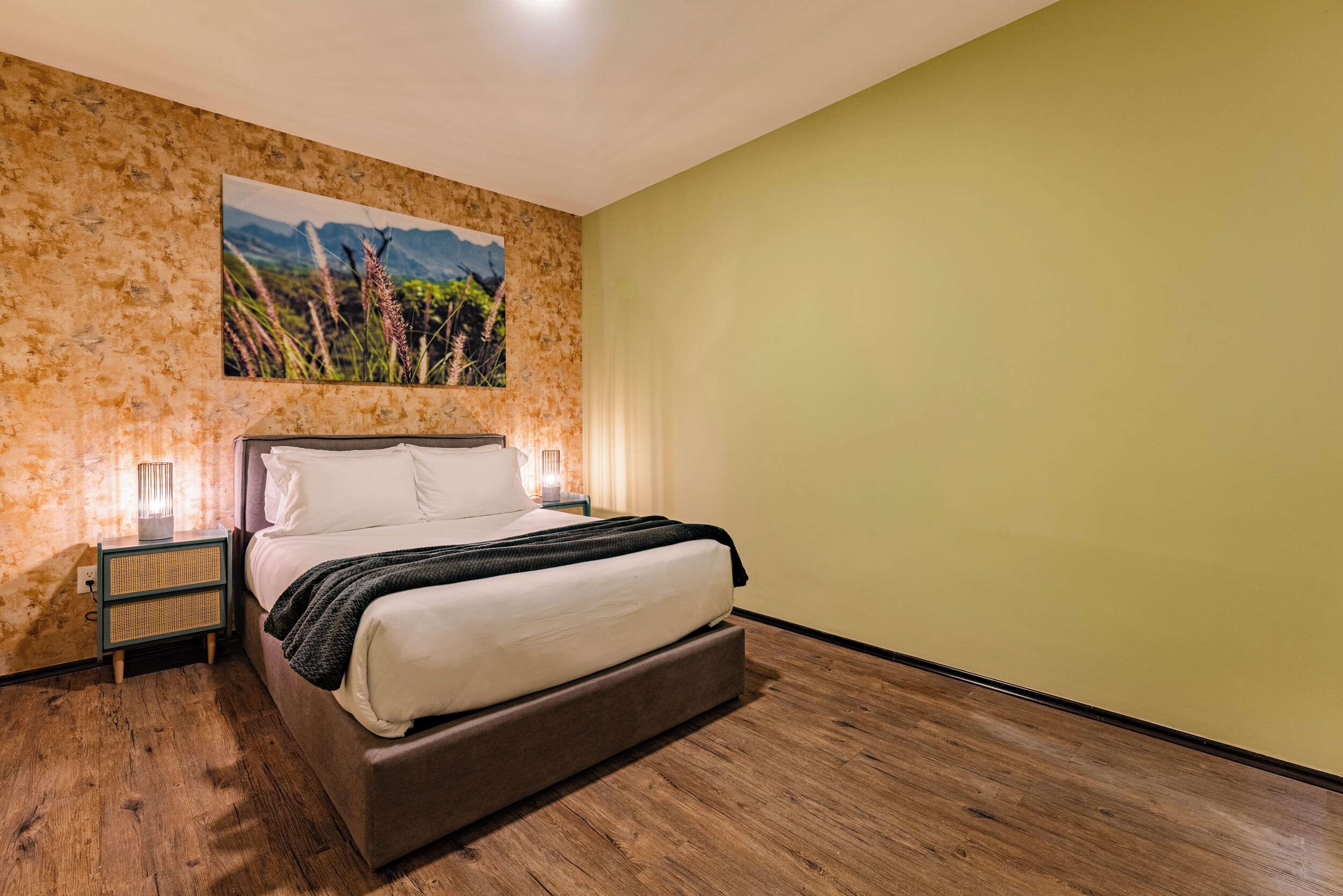 A bedroom with a bed, a nightstand, a wall-mounted lamp, and a painting of a landscape.