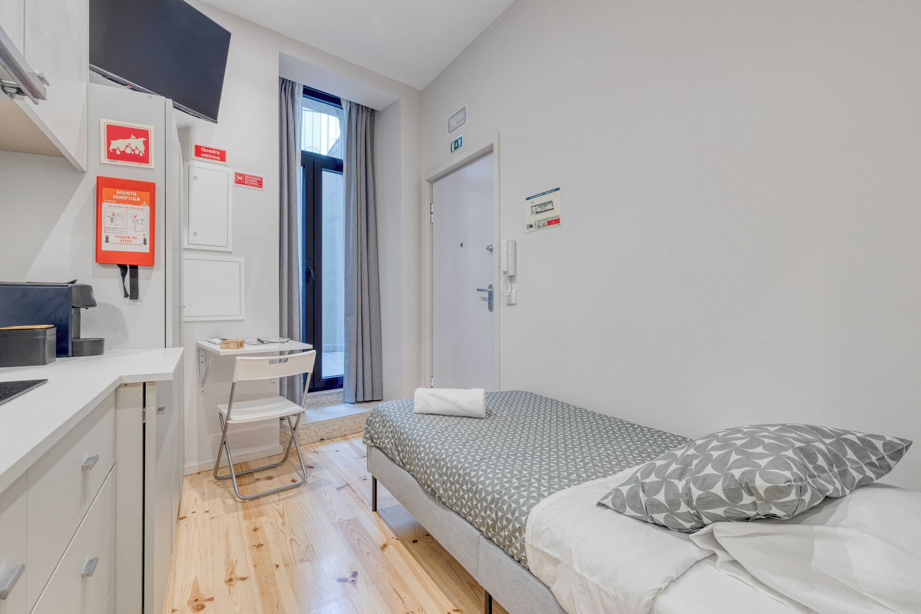 A small, clean room with a bed, a desk, and a chair. There is a door with a sign above it, a window with curtains, and a wall-mounted light switch.