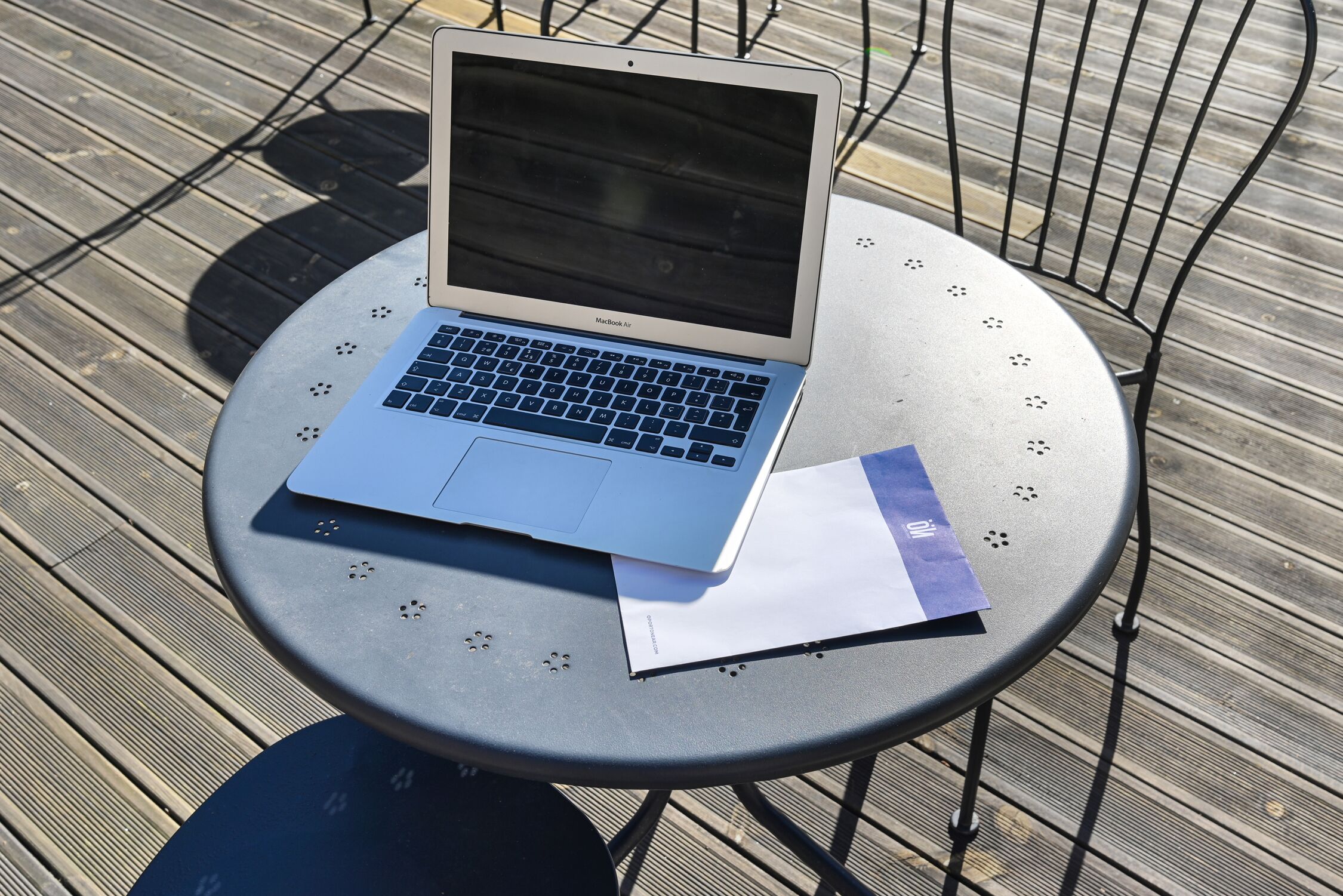 A MacBook Air on a round metal table with a chair, on a wooden deck.