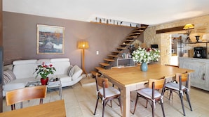 A dining area with a wooden table and chairs, a sofa, a staircase, and a kitchen area with a stone wall. - Guest Room “Aubergine” with Shared Pool, Near Sarlat (Proissans)