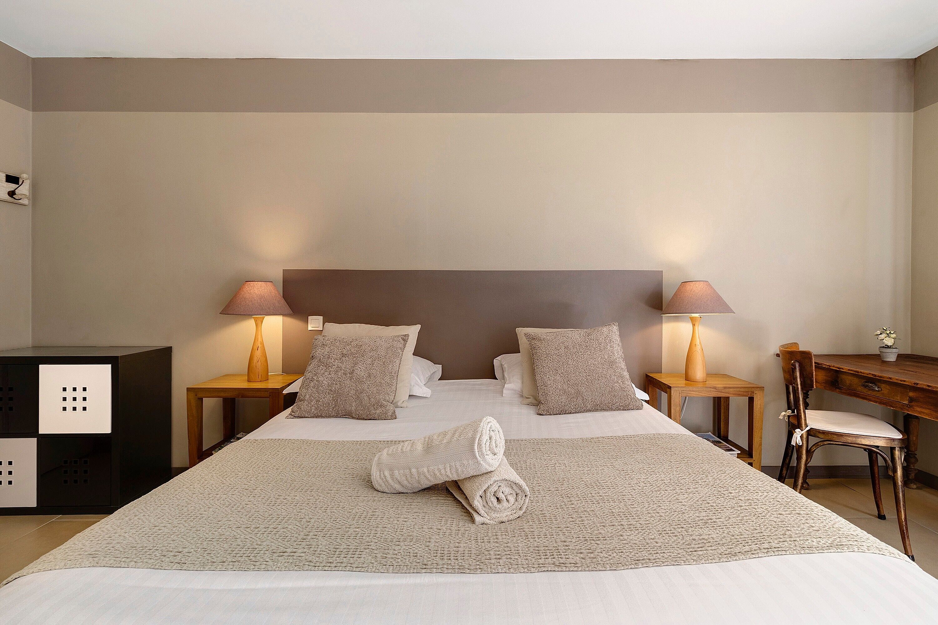 A neatly made bed with pillows, a rolled towel, and a wooden nightstand with a lamp.