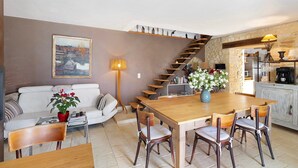 Un espace repas comprenant une table et des chaises en bois, un canapé, un escalier et un coin cuisine avec un mur en pierre.