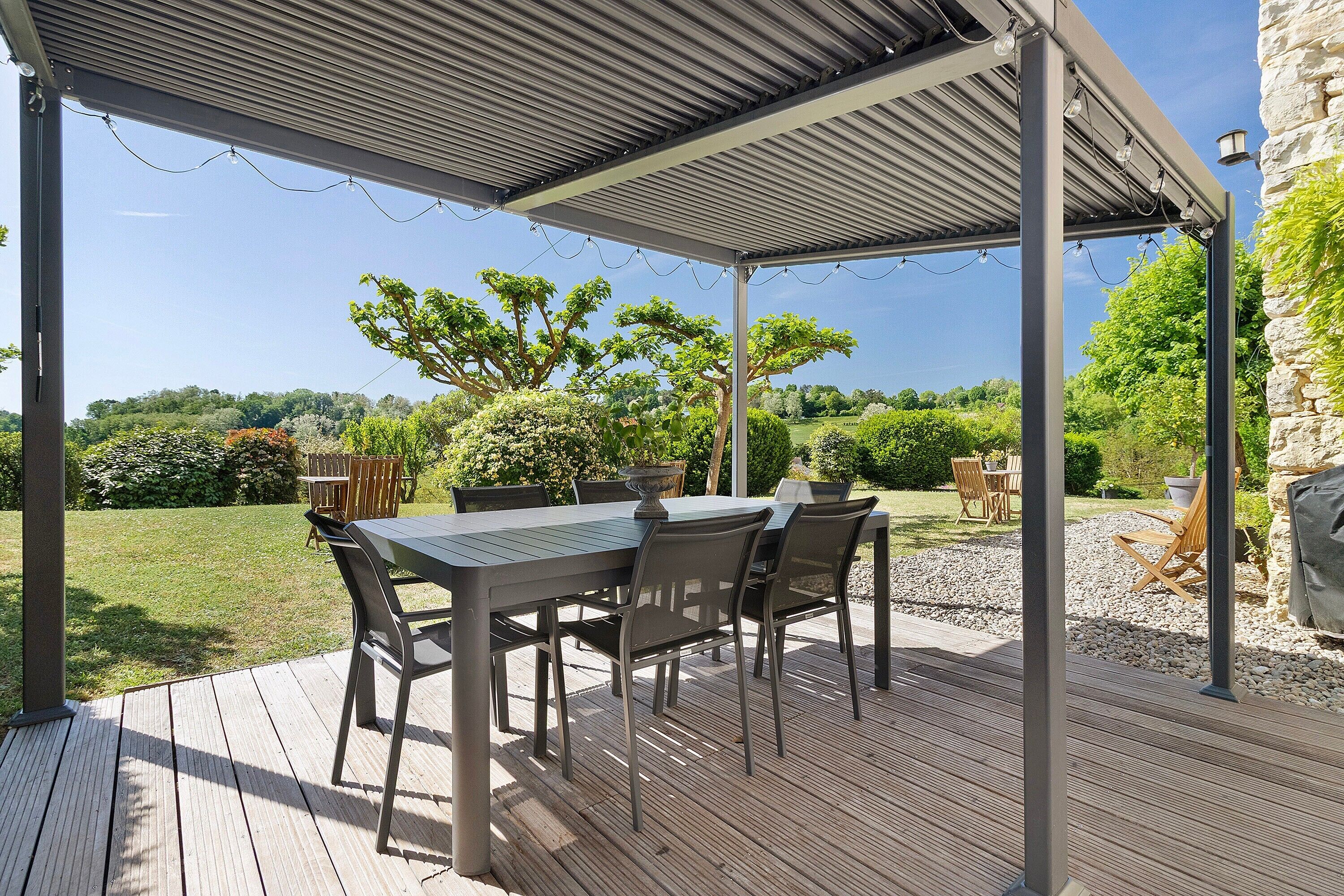 Une terrasse extérieure avec une table et des chaises abritées, entourée d’un jardin.