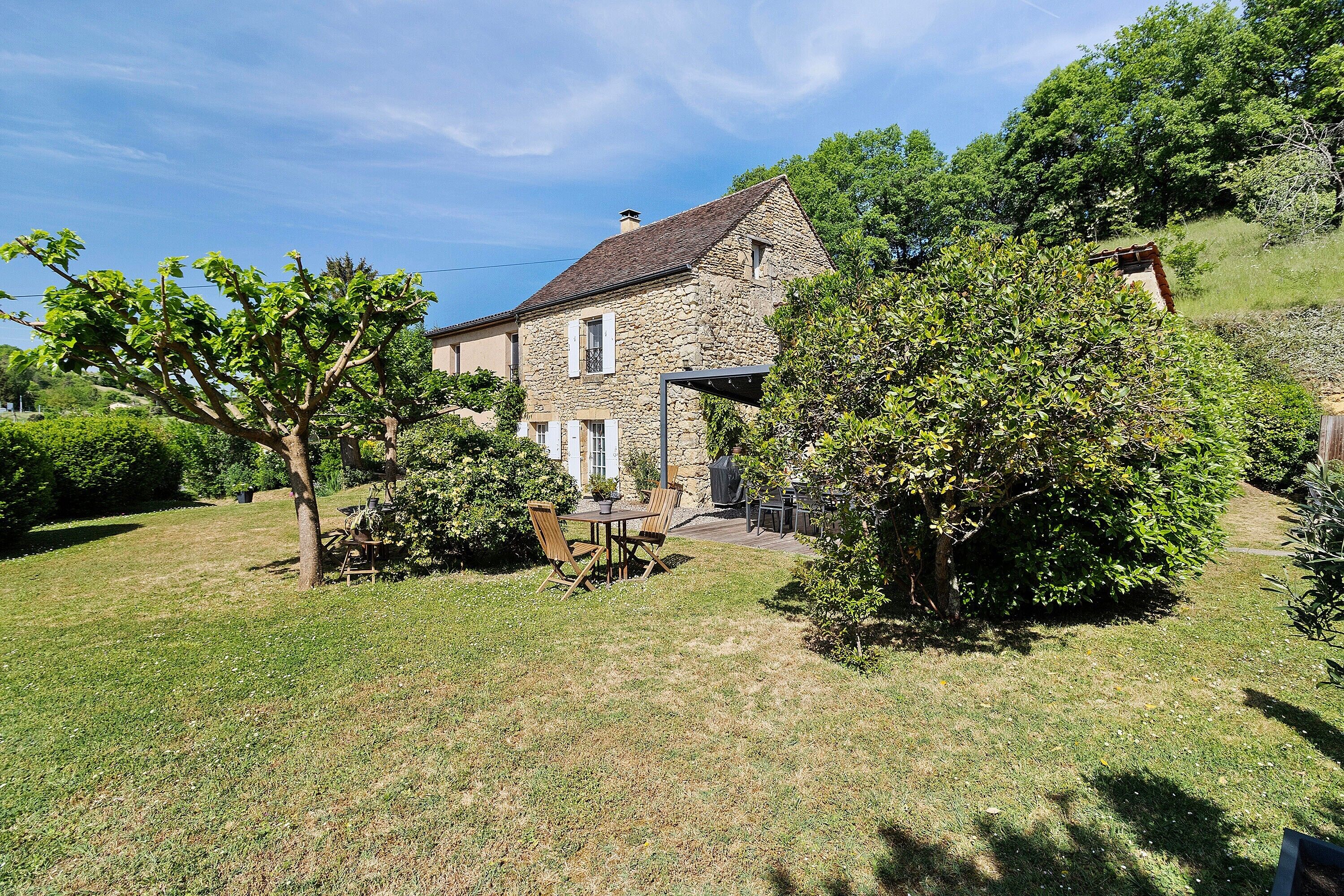 Une maison en pierre avec un jardin, une terrasse avec des chaises et une table, et un arbre au premier plan.