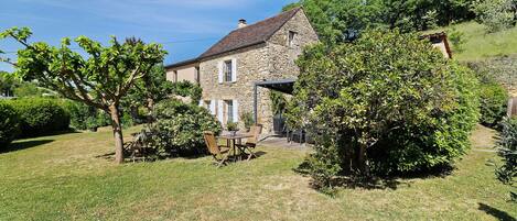 Une maison en pierre avec un jardin, une terrasse avec des chaises et une table, et un arbre au premier plan.