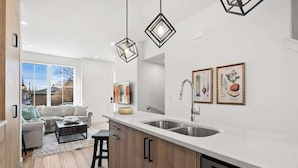 Cozinha moderna com bancada branca, armários de madeira, lava-louças e iluminação pendente.