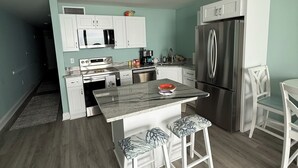 Een moderne keuken met witte kasten, een roestvrijstalen koelkast, een magnetron en een koffiezetapparaat. Er staan twee witte barkrukken met gestreept kussen.