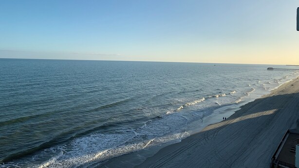 Een strand met een pier die het water in loopt, een heldere lucht en een zandstrand.