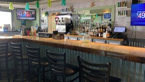 Een bar met een toonbank, barstoelen, een televisiescherm en diverse flessen en blikjes op planken.