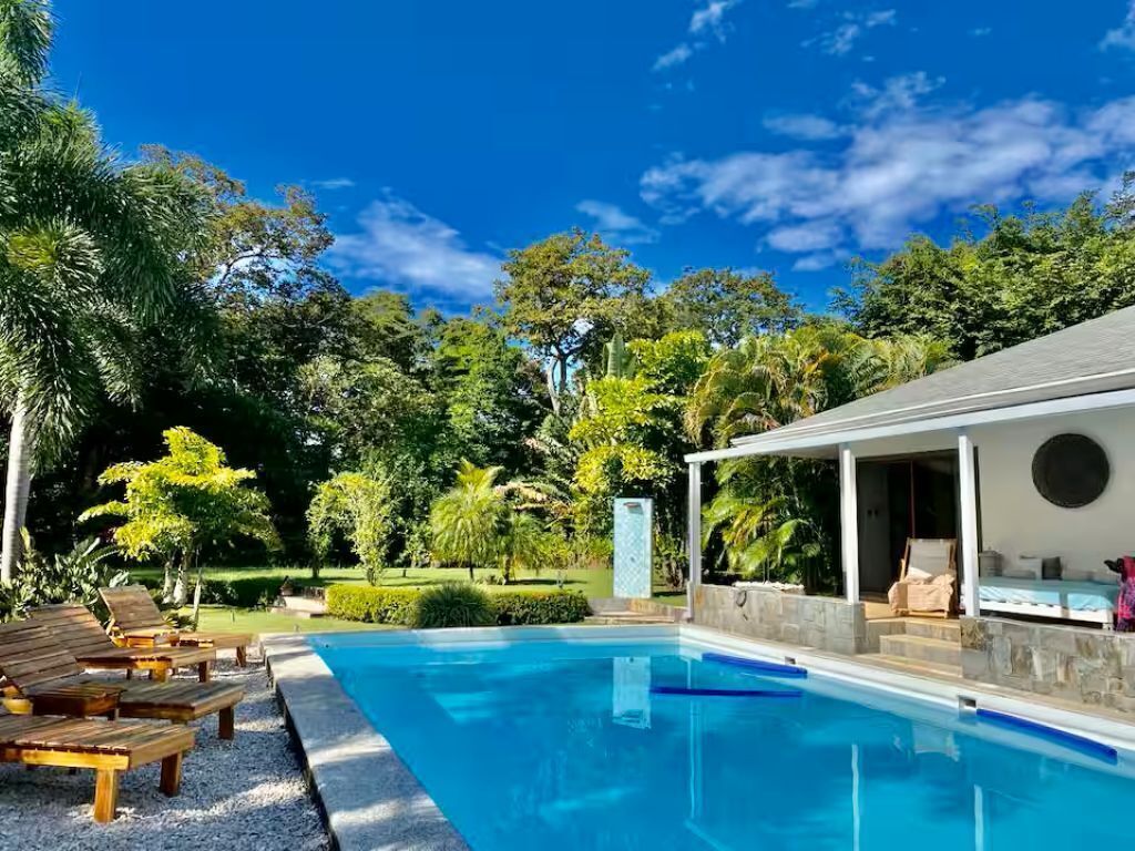Une piscine entourée de chaises longues, une terrasse couverte et une végétation luxuriante en arrière-plan.