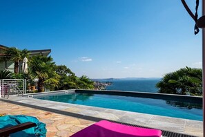 Une piscine avec vue sur la mer, entourée de palmiers et dotée d’une terrasse en pierre.