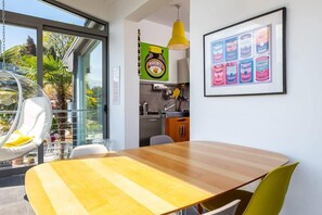 Une cuisine moderne avec une table et des chaises en bois, un luminaire suspendu et un tableau encadré représentant des conserves accroché au mur.