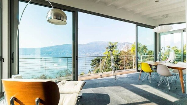 Ein modernes Zimmer mit großen Fenstern, einem Holzstuhl, einem Esstisch mit Stühlen und einer hängenden Leuchte.