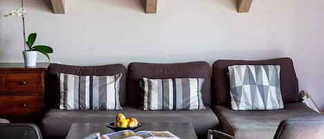 Una sala de estar moderna con un conjunto de sofá, una mesa de centro con un libro y fruta, y una planta en maceta.
