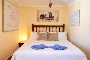 A hotel room with a bed, pillows, a painting of a scooter, and a nightstand with a lamp. - Casa Del Panadero - Stunning Views & Sunset (Frigiliana)