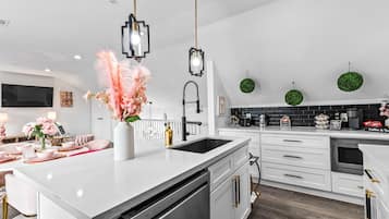 Una cocina moderna con armarios blancos, una isla central, iluminación colgante y plantas decorativas.