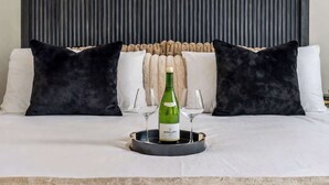 Una cama con cabecera negra, almohadas blancas y negras, y una bandeja con una botella de vino y copas.
