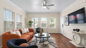 Una sala de estar con sofá, sillón, mesa de centro, televisor y ventanas.
