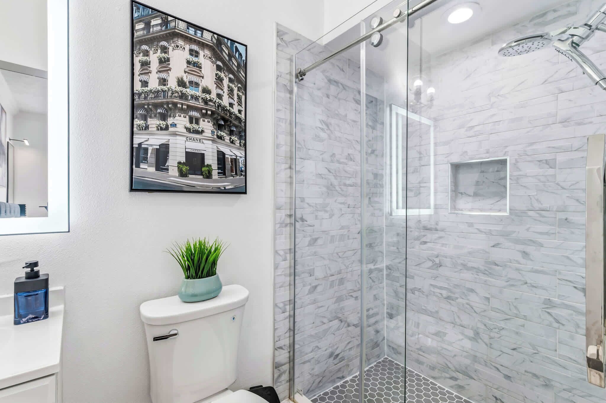 A modern bathroom with a glass shower enclosure, marble walls, and a framed picture of a building.
