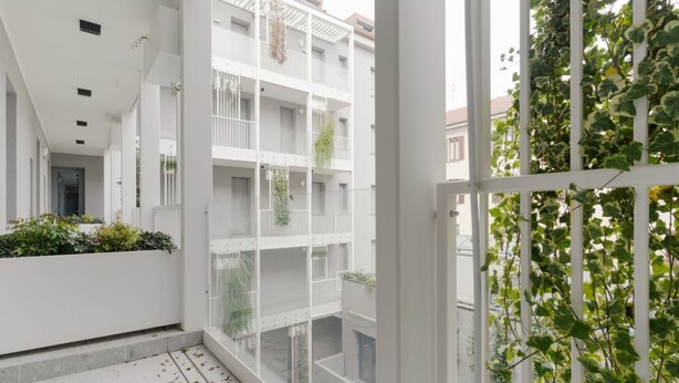 En modern byggnad med vita balkonger, vissa med växter, och utsikt över en gata med grönområden.