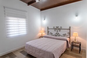 A bedroom with a bed, two bedside lamps, a window with blinds, and a wooden ceiling.