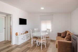 A modern living room with a round dining table, white chairs, a sofa, a flat-screen TV, and a window with blinds.