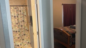 A room with a wooden floor, a bed with a plaid blanket, a shower with a cactus-patterned curtain, and a light fixture on the ceiling. - Casa Azul - Home w/fenced yard (Buda)