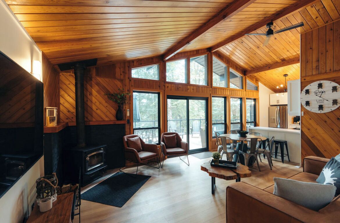A cozy cabin interior with a fireplace, wooden walls, large windows, and a dining area.