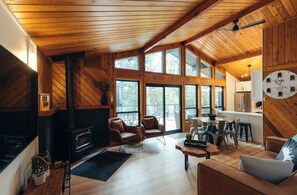 A cozy cabin interior with a fireplace, wooden walls, large windows, and a dining area. - The Cedars Haliburton: Cedars East - lakeside cottage with sauna (Dysart Et Al)