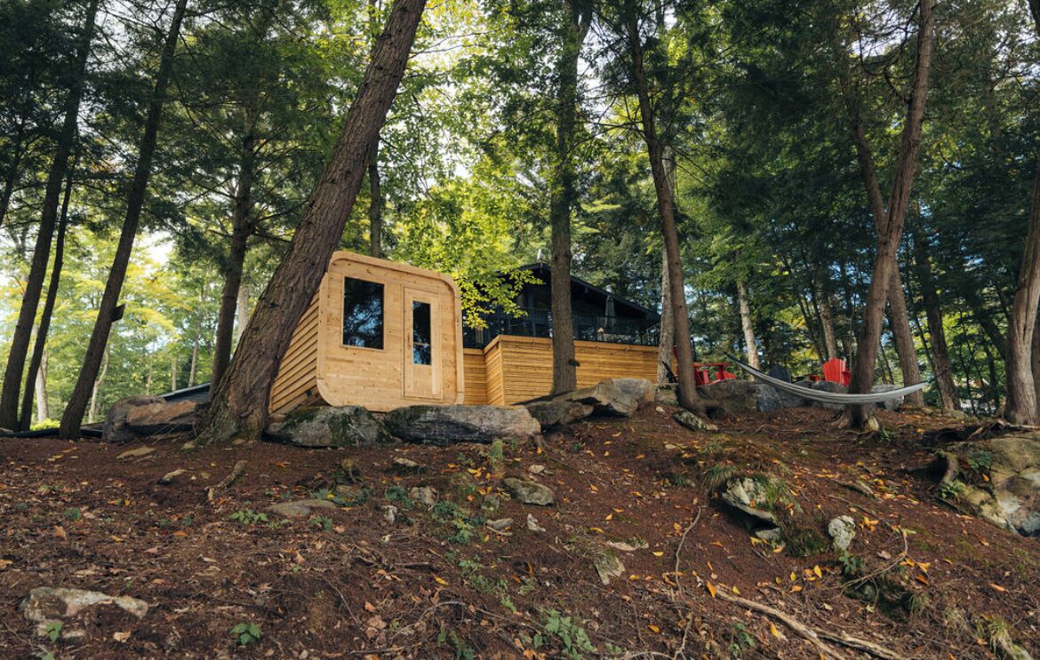 A wooden cabin in a forest with a hammock and a red chair.
