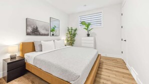 Ein Schlafzimmer mit Bett, Nachttisch, Wandspiegel, gerahmtem Bild, Topfpflanzen und einem Fenster mit Jalousien.