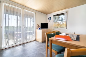 Una habitación con techo de madera, una mesa de comedor con sillas, un televisor sobre un mueble bajo y un ventanal con vistas a árboles.