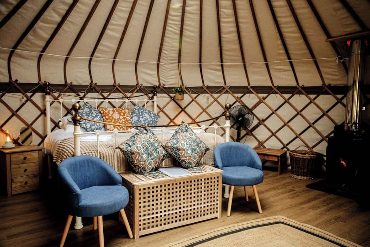 A cozy interior of a yurt with a bed, two blue chairs, a small table, and a heater.