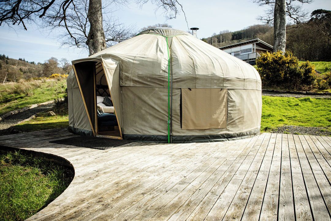 A round, beige tent-like structure with an open entrance, situated on a wooden deck with a grassy area in the background.