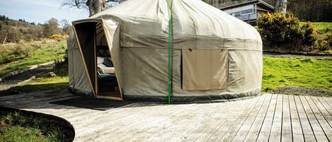 A round, beige tent-like structure with an open entrance, situated on a wooden deck with a grassy area in the background.
