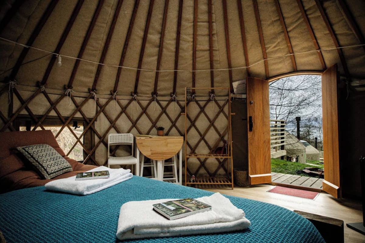 A yurt interior with a bed, a small table, a chair, and a view of the outside.