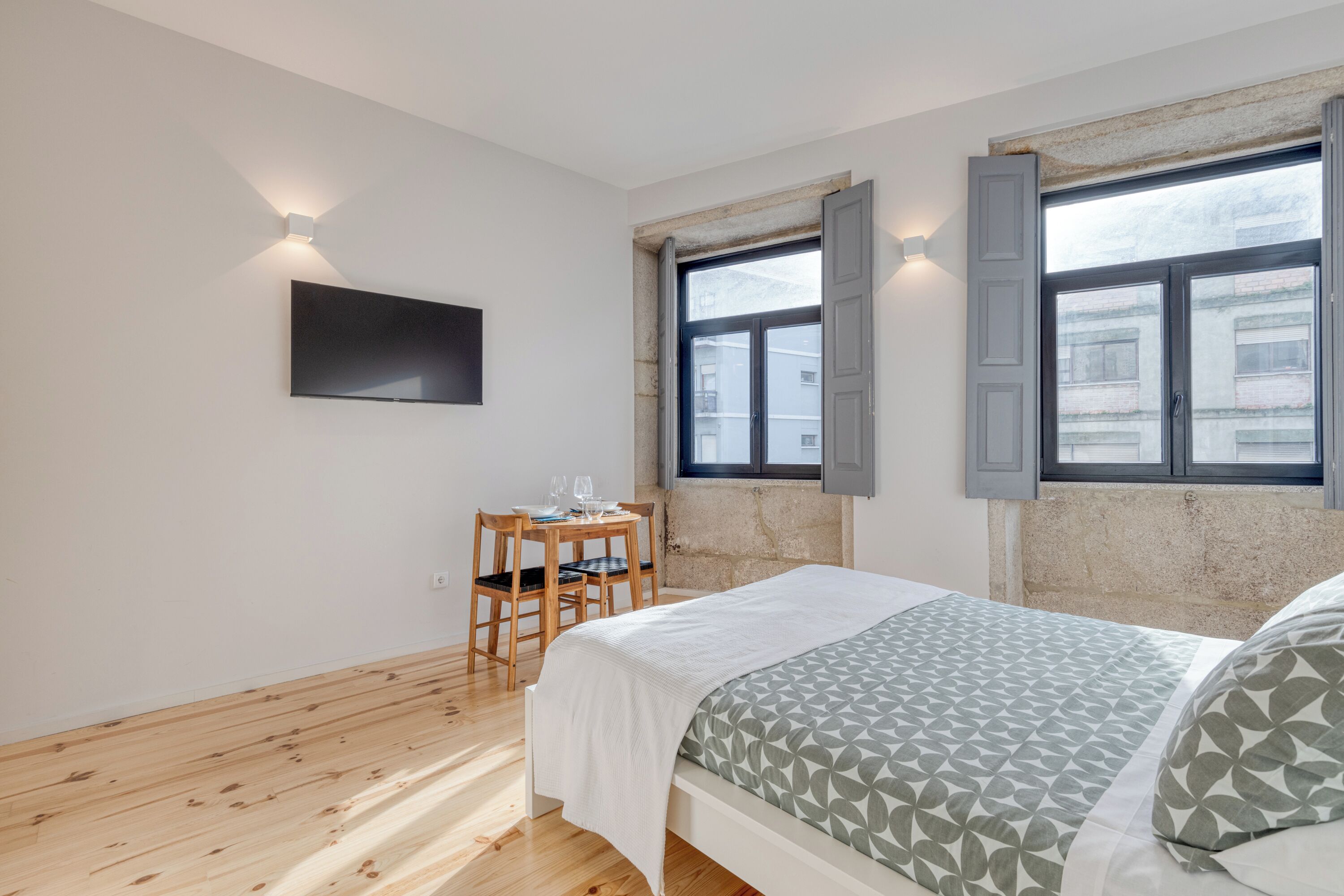 A bedroom with a bed, a TV mounted on the wall, a window with shutters, and a small table with a chair.
