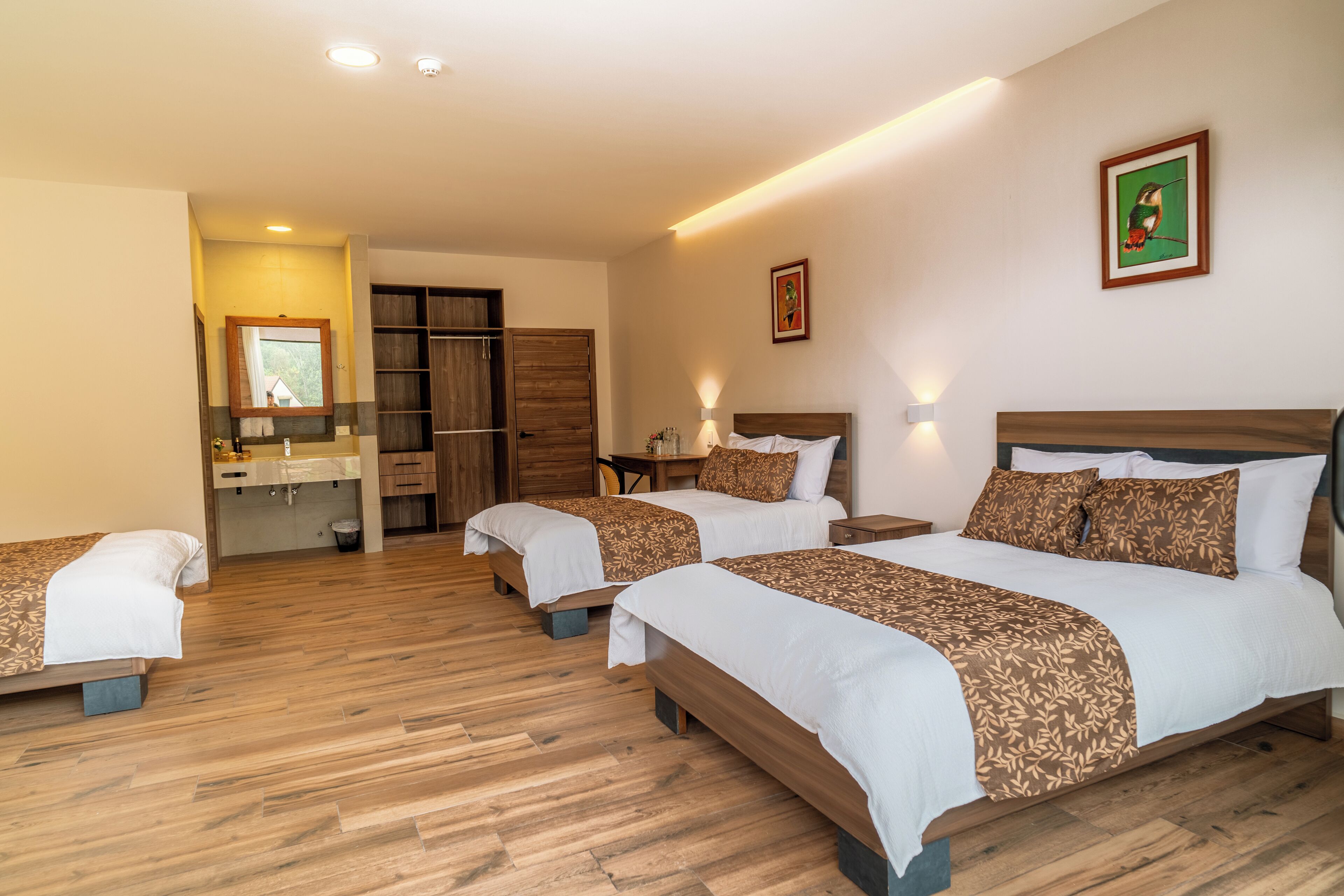 A hotel room with three beds, a wooden floor, a bathroom with a sink and mirror, and a door leading to a closet.
