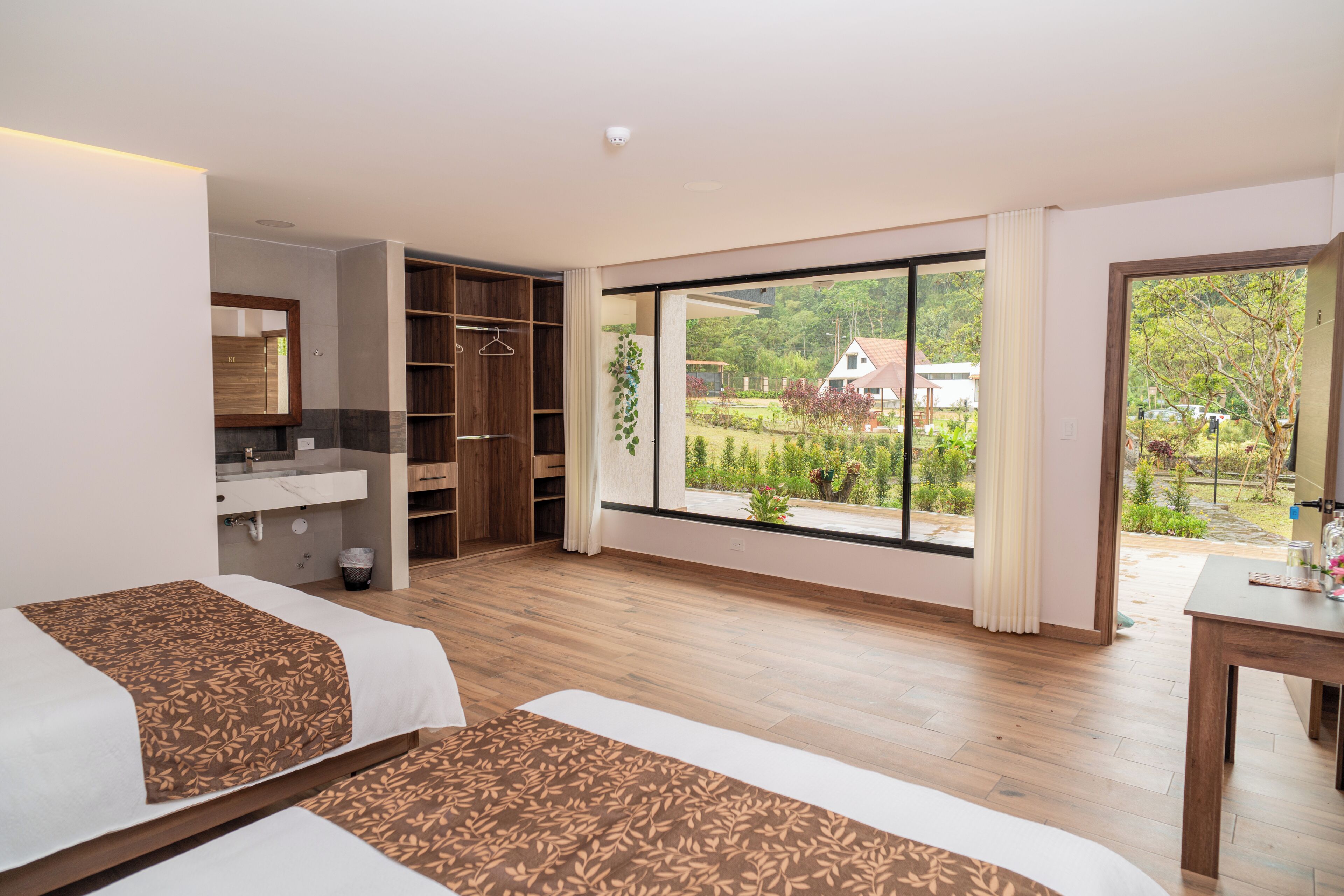 Una habitación de hotel moderna con un ventanal grande, suelo de madera, cama, escritorio y baño con espejo y lavabo.