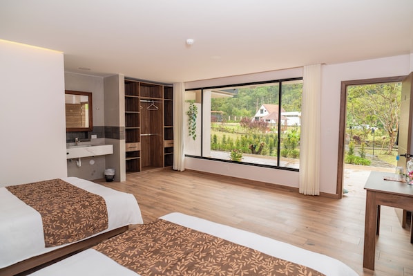 A modern hotel room with a large window, a wooden floor, a bed, a desk, and a bathroom with a mirror and sink. - Hostería Pilpikuna (Mindo)