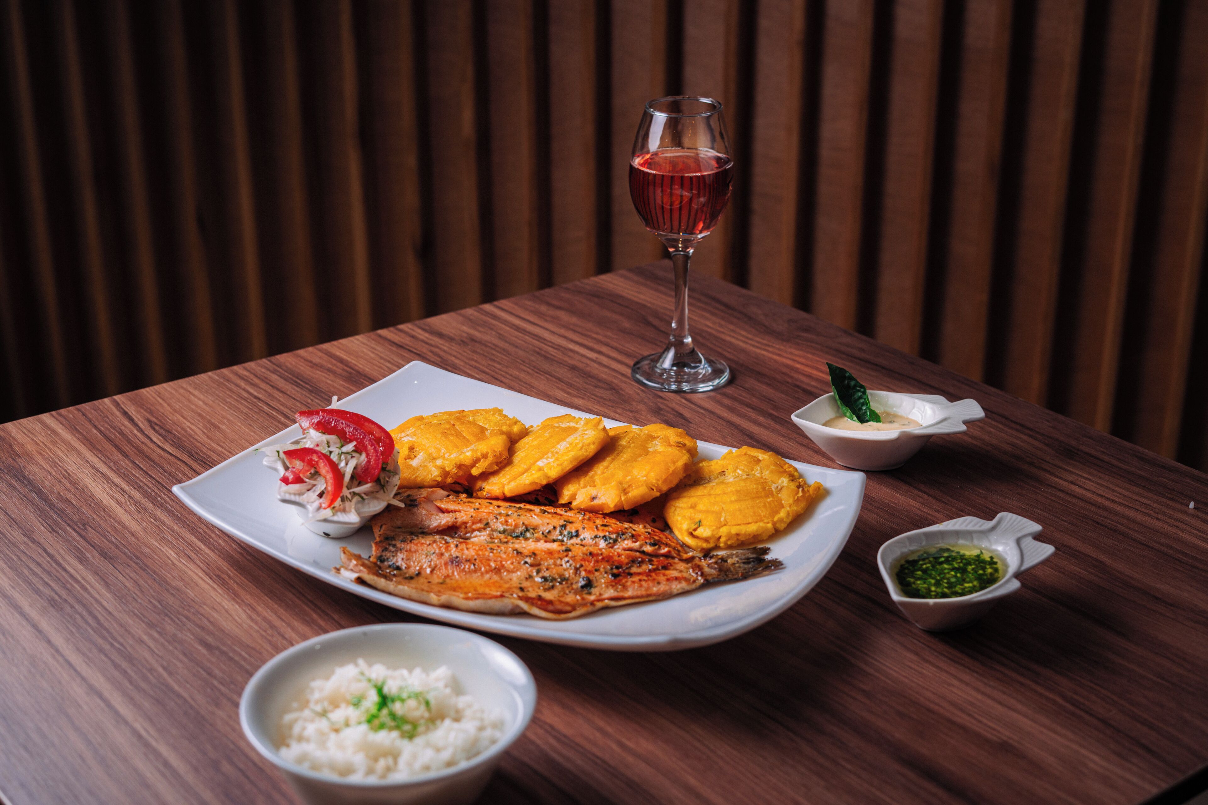 A wooden table with a plate of food, a glass of red wine, and small bowls of condiments.