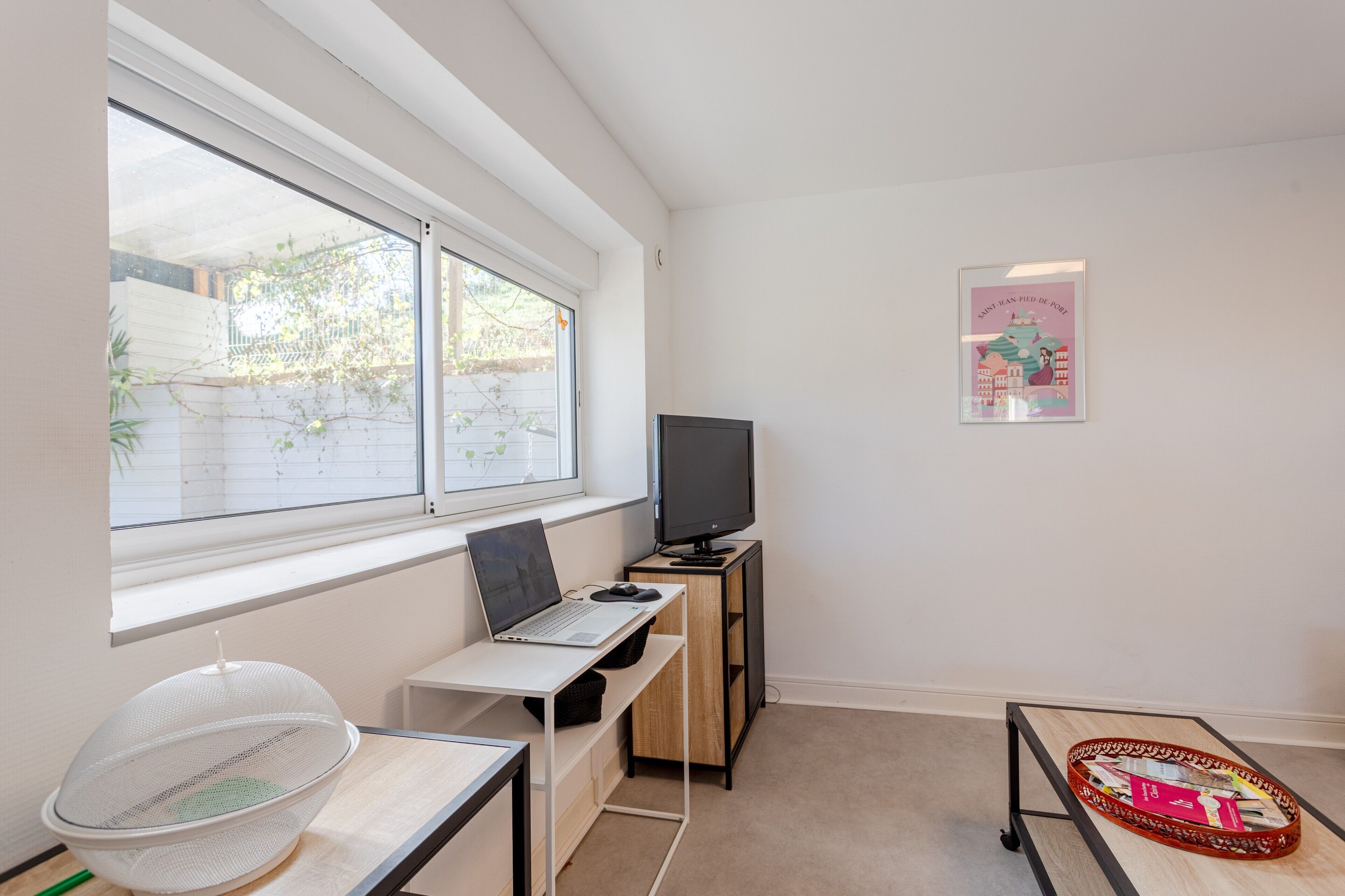 Una habitación con escritorio, laptop, televisión, ventana con vistas a la vegetación y un cartel en la pared.